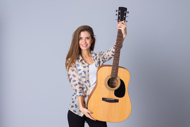 Giovane donna sorridente in camicetta beige e pantaloni neri che tiene una chitarra su sfondo grigio in studio