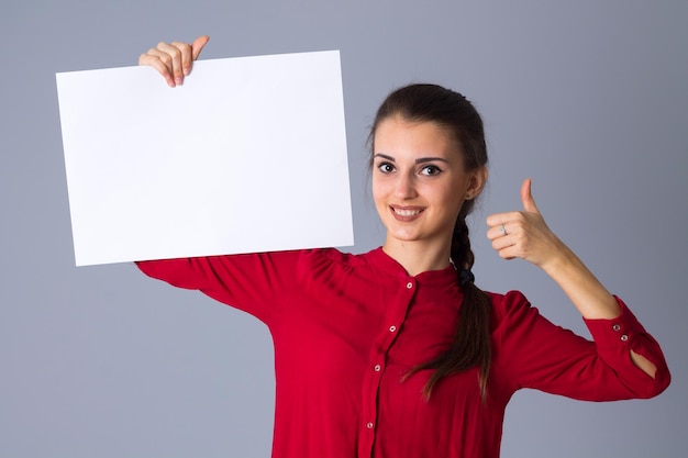 Giovane donna sorridente in camicetta rossa che tiene foglio di carta bianco e mostra il pollice in su su sfondo grigio in studio