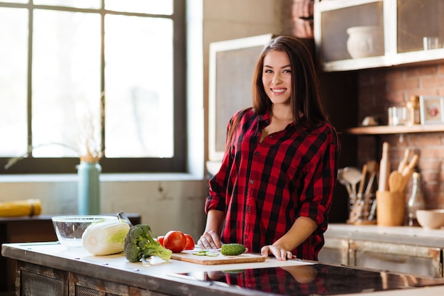 Giovane donna sorridente in camicia rossa in piedi al tavolo in cucina