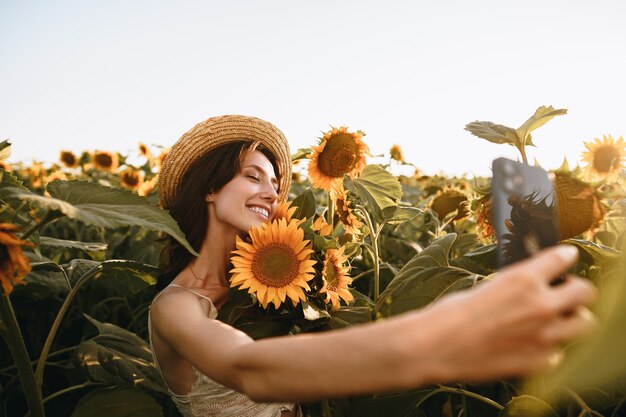 Giovane donna sorridente in cappello che prende la foto del selfie nel campo del girasole