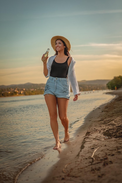 Giovane donna sorridente in cappello di paglia che si rilassa al tramonto sulla riva del fiume. Sta camminando lungo la spiaggia del fiume e si diverte.
