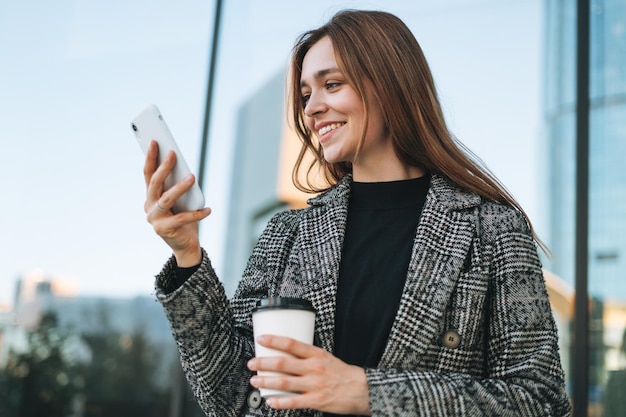 Giovane donna sorridente in cappotto con tazza di caffè utilizzando il telefono cellulare in via della città di sera