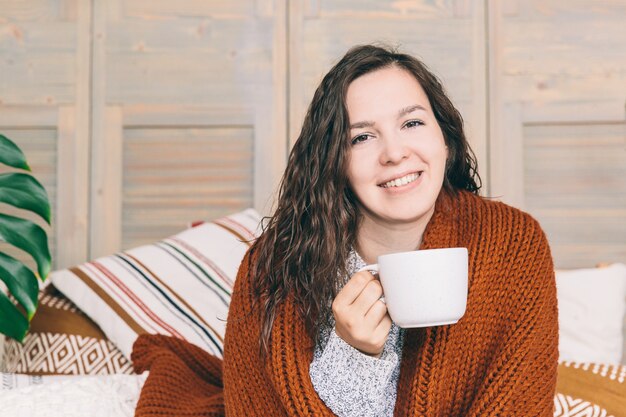 Giovane donna sorridente in coperta marrone sdraiato sul letto con tazza bianca.