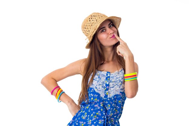 Giovane donna sorridente in maglietta blu e pantaloncini gialli con braccialetti colorati e cappello che si toccano le labbra su sfondo bianco in studio