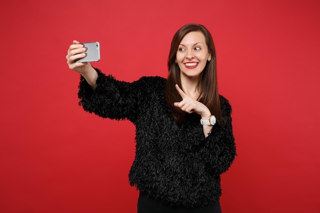 Giovane donna sorridente in maglione di pelliccia nera che punta il dito indice facendo selfie sparato sul telefono cellulare isolato su sfondo rosso brillante della parete. Persone sincere emozioni, concetto di stile di vita. Mock up copia spazio.
