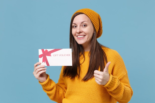 Giovane donna sorridente in maglione giallo e cappello in posa isolata su sfondo blu ritratto in studio. Persone sincere emozioni concetto di stile di vita. Mock up spazio di copia. Mostrando il pollice in su tenere il buono regalo.