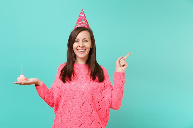 Giovane donna sorridente in maglione rosa lavorato a maglia, cappello di compleanno che indica il dito indice da parte tenere in mano la torta con la candela isolata su priorità bassa blu in studio. Concetto di stile di vita della gente. Mock up copia spazio.