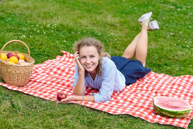 Giovane donna sorridente rilassante all'aperto e fare un picnic è sdraiata su una coperta di globuli rossi sull'erba nel prato