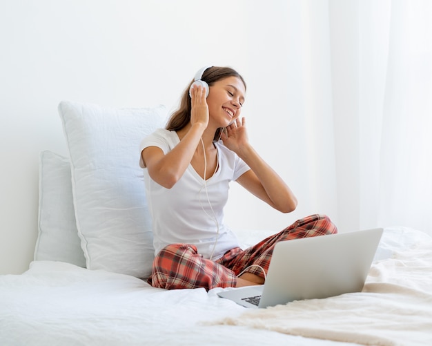 Giovane donna sorridente seduta sul letto in camera da letto e ascoltare musica o guardare film