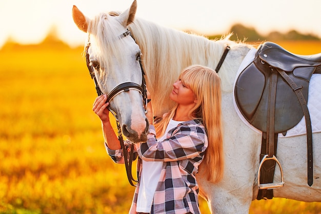 Giovane donna sorridente soddisfatta allegra felice sveglia che abbraccia e che accarezza il bellissimo cavallo bianco al prato al tramonto all'ora d'oro