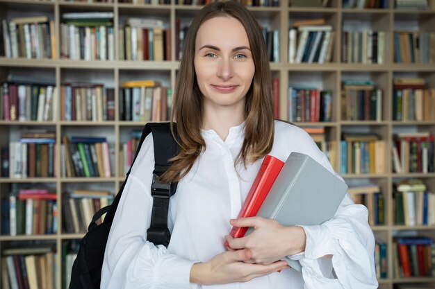 Giovane donna sorridente sta con libri e uno zaino in biblioteca