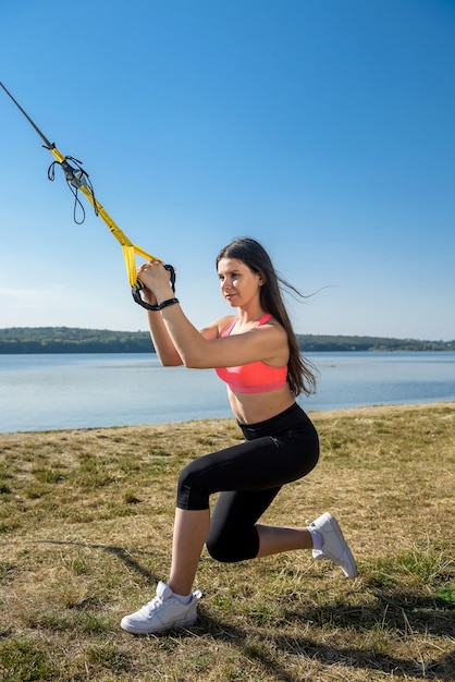 Giovane donna sottile facendo allenamento in sospensione con cinghie di fitness all'aperto vicino al lago durante il giorno