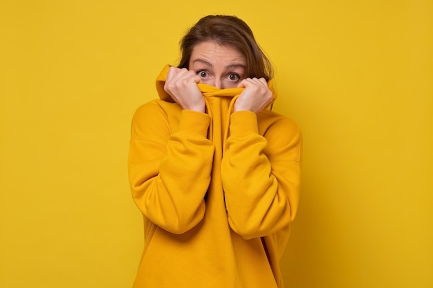 Giovane donna spaventata, impaurita e ansiosa, nascondendo il viso, guardando la fotocamera su uno sfondo giallo.