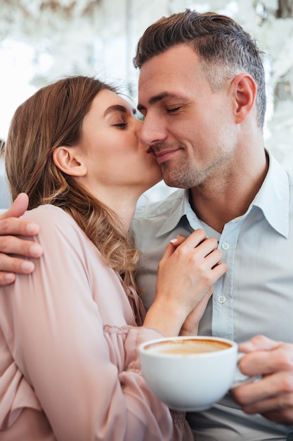 Giovane donna splendida che abbraccia e che bacia il suo uomo maschile del ragazzo nell'accogliente caffè della città, mentre uomo soddisfatto che beve tè o caffè