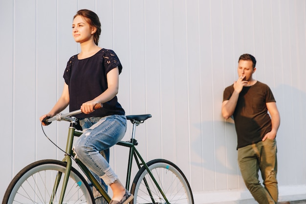 Giovane donna splendida che guida una bicicletta, mentre ragazzo bello alla moda che fuma e che la guarda con ammirazione. All'aperto.