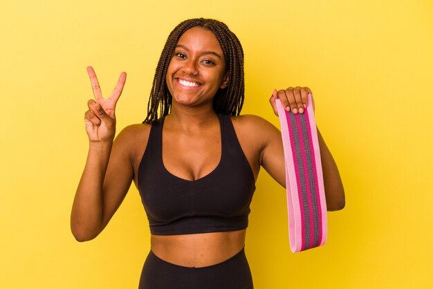 Giovane donna sportiva afroamericana che tiene una fascia elastica isolata su sfondo giallo che mostra il numero due con le dita.