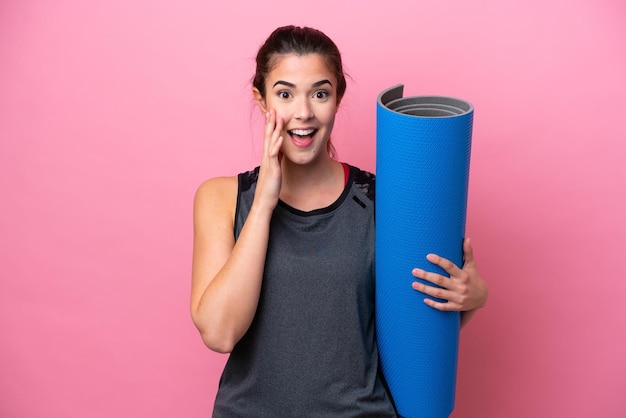 Giovane donna sportiva brasiliana che va a lezioni di yoga mentre tiene un tappetino isolato su sfondo rosa con espressione facciale sorpresa e scioccata