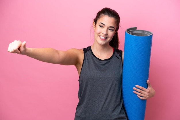 Giovane donna sportiva brasiliana che va alle lezioni di yoga mentre tiene un tappetino isolato su sfondo rosa che dà un gesto del pollice in alto