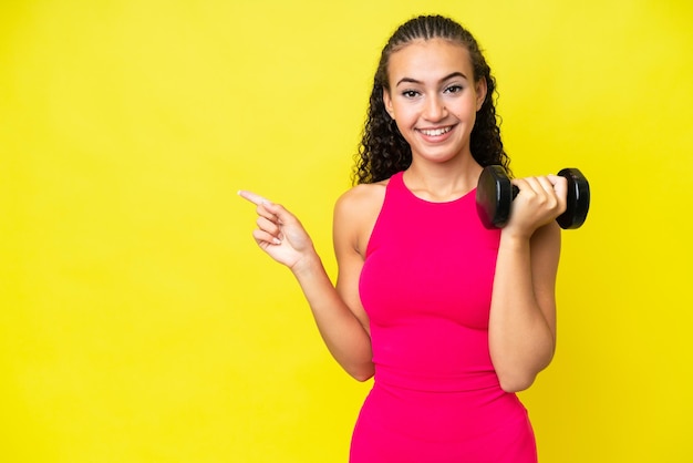 Giovane donna sportiva che fa sollevamento pesi isolato su sfondo giallo che punta il dito di lato