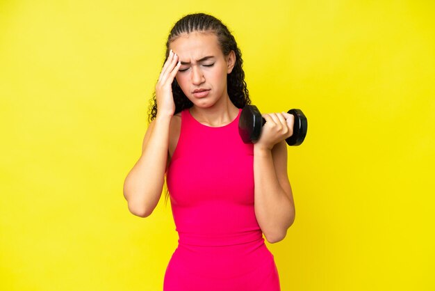 Giovane donna sportiva che fa sollevamento pesi isolato su sfondo giallo con mal di testa