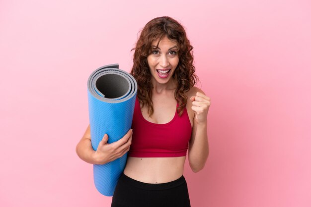 Giovane donna sportiva che va a lezioni di yoga mentre tiene un tappetino isolato su sfondo rosa che celebra una vittoria in posizione di vincitore