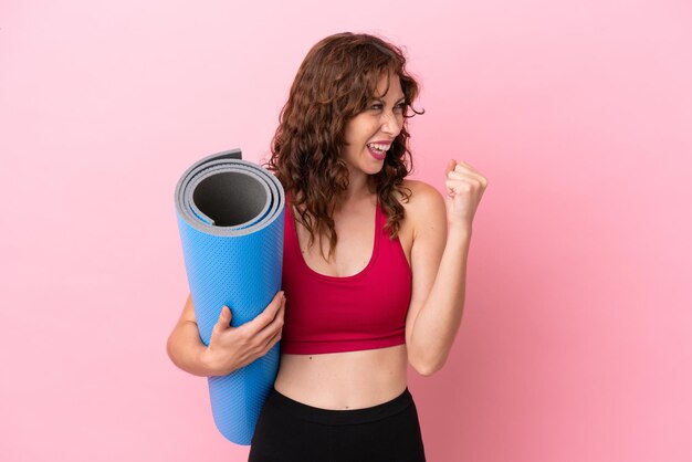 Giovane donna sportiva che va a lezioni di yoga mentre tiene un tappetino isolato su sfondo rosa che celebra una vittoria