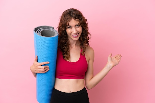 Giovane donna sportiva che va a lezioni di yoga mentre tiene un tappetino isolato su sfondo rosa che estende le mani di lato per invitare a venire