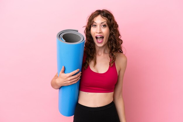 Giovane donna sportiva che va a lezioni di yoga mentre tiene un tappetino isolato su sfondo rosa con espressione facciale a sorpresa