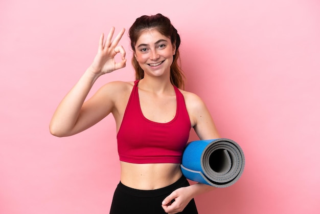 Giovane donna sportiva che va alle lezioni di yoga tenendo in mano un tappetino che mostra il segno ok con le dita