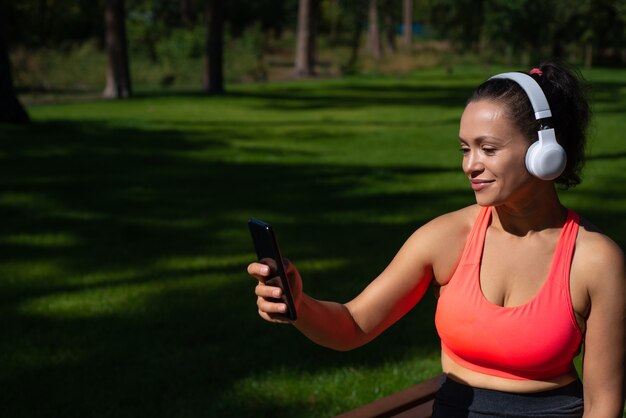 Giovane donna sportiva con le cuffie che tengono smartphone e playlist cheking mentre si ascolta la musica nel parco cittadino