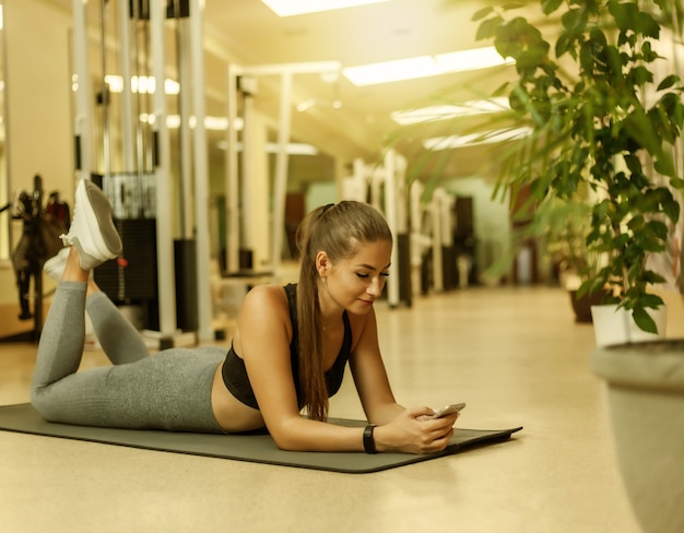 Giovane donna sportiva in abbigliamento sportivo che utilizza smartphone mentre si trova sul tappetino in palestra. Pausa allenamento. Concetto di stile di vita sano