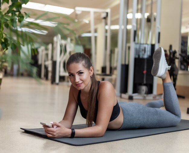 Giovane donna sportiva in abiti sportivi utilizzando smartphone mentre giace sul tappetino in palestra. Pausa allenamento. Concetto di stile di vita sano