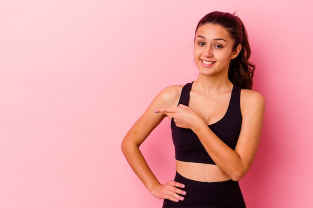 Giovane donna sportiva isolata sul muro rosa sorridendo e indicando da parte, mostrando qualcosa in uno spazio vuoto