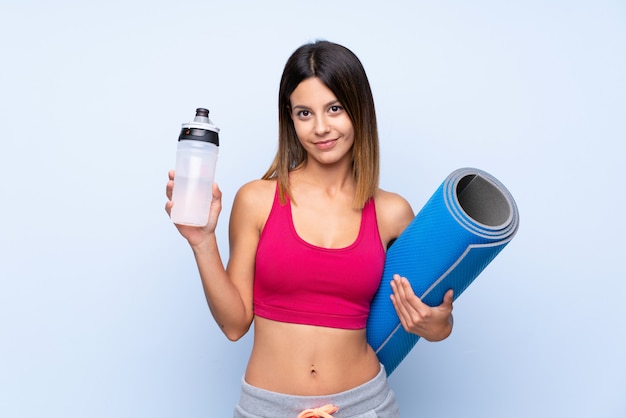 Giovane donna sportiva sopra la parete blu isolata con la bottiglia di acqua di sport e con una stuoia