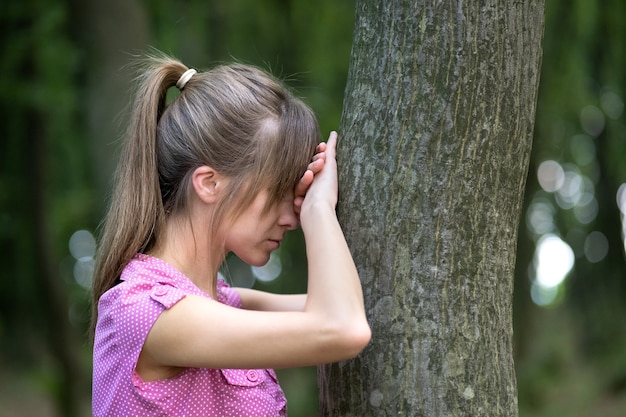 Giovane donna stanca che si appoggia al tronco di albero nella foresta di estate.