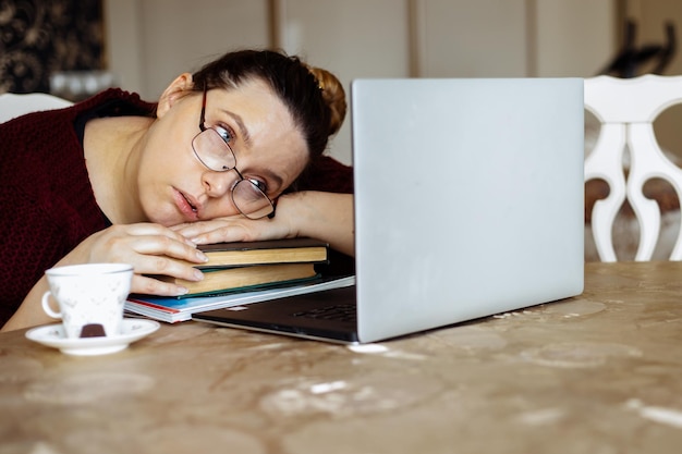 Giovane donna stanca con gli occhiali sdraiata sui libri e guardando lo schermo del laptop alla scrivania nella stanza di casa t