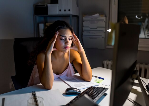 Giovane donna stanca di affari con l&#39;emicrania che si siede al computer nel posto di lavoro - straordinari di notte