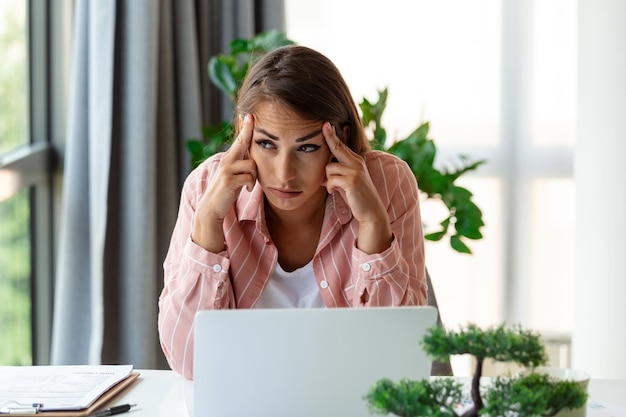 Giovane donna stanca esausta dal lavoro di studio Annoiata e frustrata guardando il laptop Testa appoggiata a portata di mano Spazio luminoso grandi finestre Concetto di casa Concetto di stress