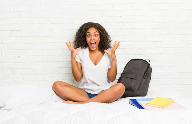 Giovane donna studentessa afroamericana sul letto che celebra una vittoria o un successo, è sorpreso e scioccato.