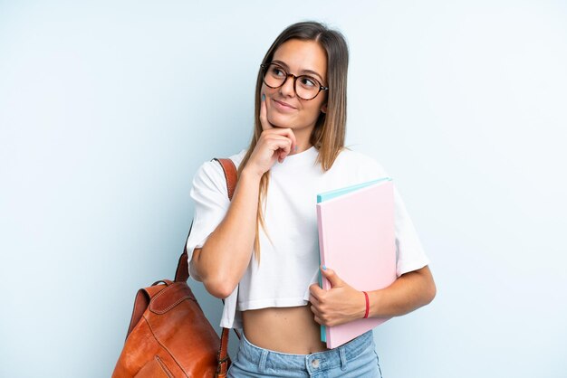 Giovane donna studentessa isolata su sfondo blu che pensa un'idea mentre guarda in alto