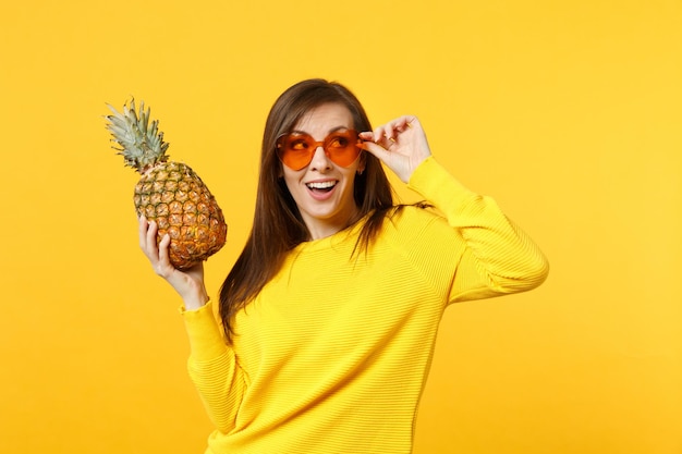 Giovane donna stupita con gli occhiali del cuore che osserva in su, tiene la frutta fresca matura dell'ananas isolata sul fondo giallo arancio della parete in studio. Persone stile di vita vivido, relax concetto di vacanza. Mock up copia spazio