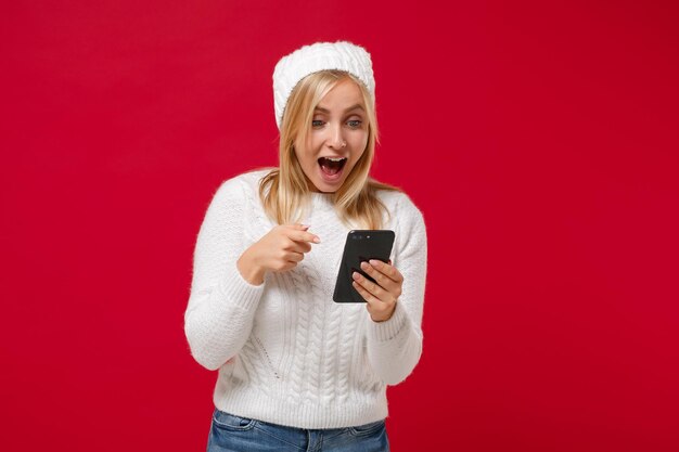 Giovane donna stupita in maglione bianco, cappello isolato su sfondo muro rosso. Stile di vita sano della moda, concetto di stagione fredda. Simula lo spazio della copia. Puntare il dito indice sul cellulare, digitare un messaggio sms.