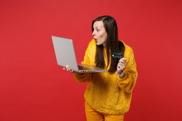 Giovane donna stupita in maglione di pelliccia gialla che tiene carta bancaria di credito, computer pc portatile isolato su sfondo rosso brillante parete in studio. Persone sincere emozioni, concetto di stile di vita. Mock up copia spazio.