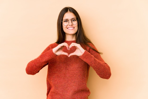 Giovane donna su beige che sorride e che mostra una forma del cuore con le mani.