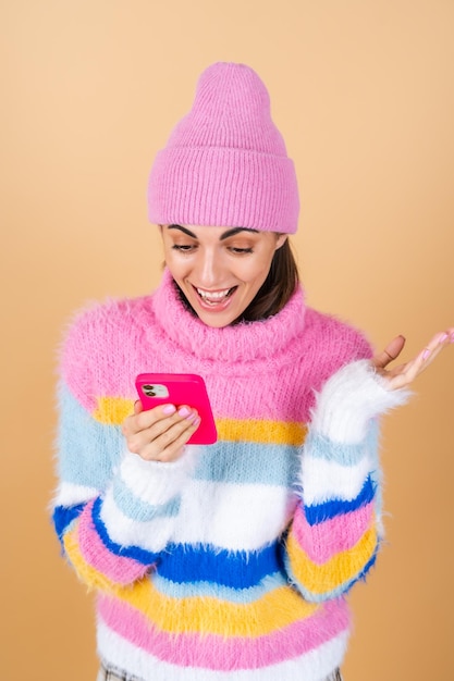 Giovane donna su beige in un maglione lavorato a maglia e un cappello con un telefono cellulare che ride con sorpresa e divertimento, leggendo messaggi, barzellette, congratulazioni