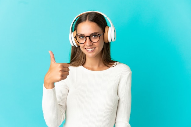 Giovane donna su sfondo blu isolato ascoltando musica e con il pollice in alto