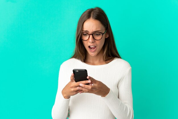 Giovane donna su sfondo blu isolato guardando la telecamera mentre si utilizza il cellulare con espressione sorpresa