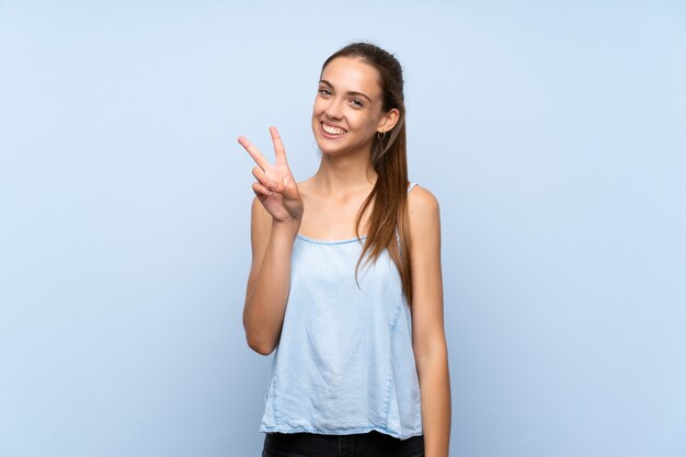 Giovane donna su sfondo blu isolato, sorridente e mostrando il segno della vittoria