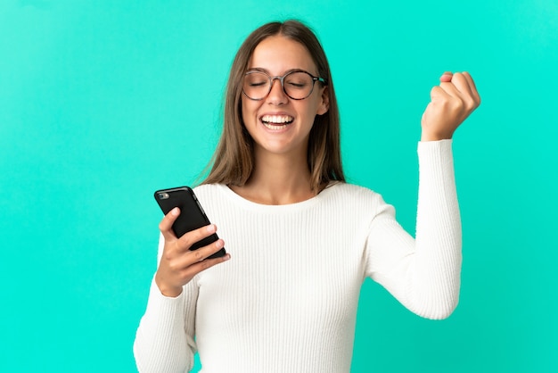 Giovane donna su sfondo blu isolato utilizzando il telefono cellulare e facendo gesto di vittoria