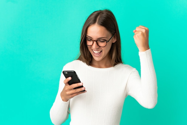 Giovane donna su sfondo blu isolato utilizzando il telefono cellulare e facendo il gesto di vittoria